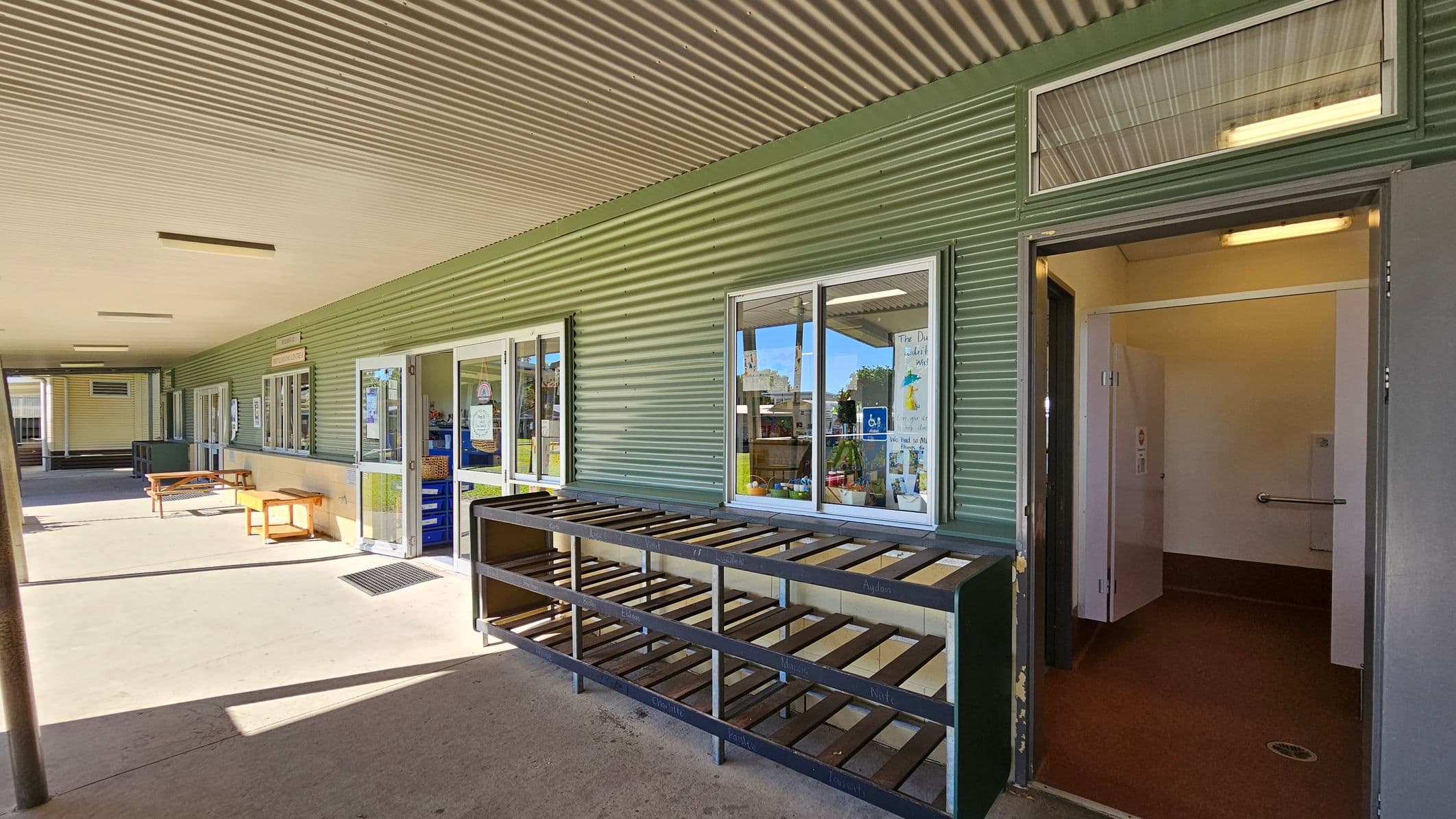A covered outdoor area with a corrugated metal ceiling and walls. On the right, there's an open door leading to a room with white walls and a handrail. In the centre, a shoe rack is positioned against the wall beneath a window displaying posters. On the left, there are several windows and a wooden bench. The floor is concrete, and the area is well-lit with ceiling lights.