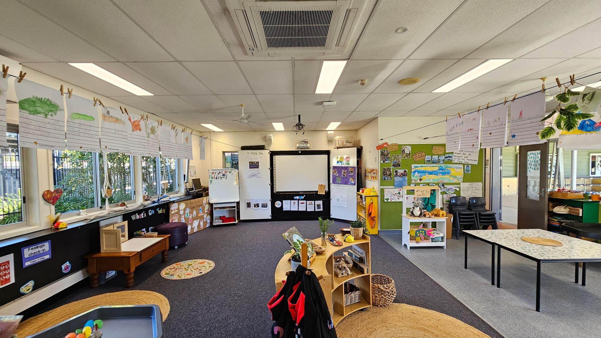 A classroom with a carpeted floor and a ceiling with fluorescent lights. On the left, there are large windows with children's artwork hanging above them. In the centre, a whiteboard and a smartboard are flanked by storage units. On the right, a green bulletin board displays more artwork, and there are stacked chairs and a small table. The foreground features a circular shelf with educational materials and a woven rug.