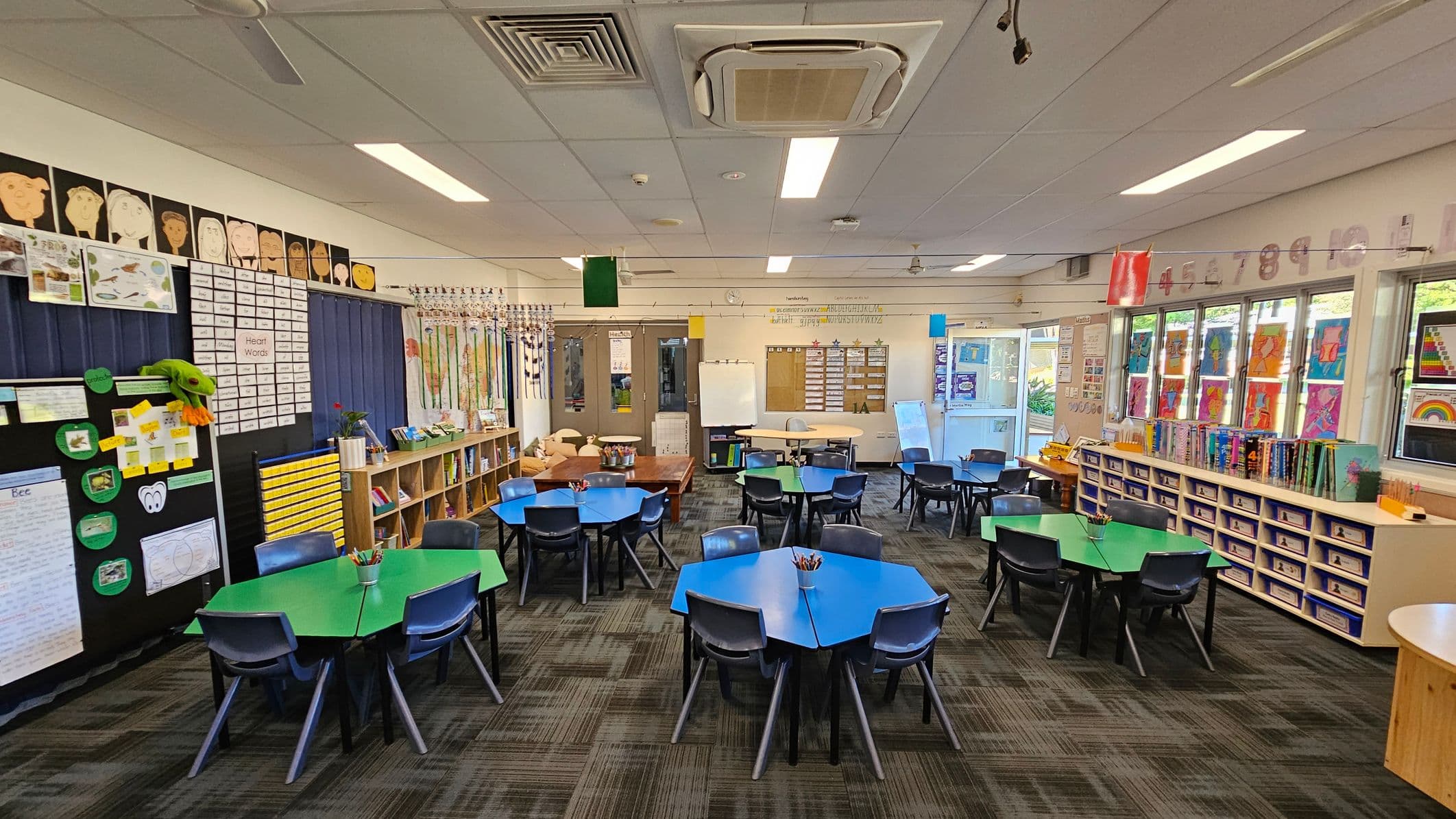 Classroom with a patterned carpet floor and a ceiling with fluorescent lights and air conditioning vents. In the foreground, hexagonal tables with blue and green tops are surrounded by grey chairs. The left wall features educational posters and a bookshelf. The right side has windows with colourful artwork and a row of storage cubbies. The back wall includes a whiteboard and more educational materials.