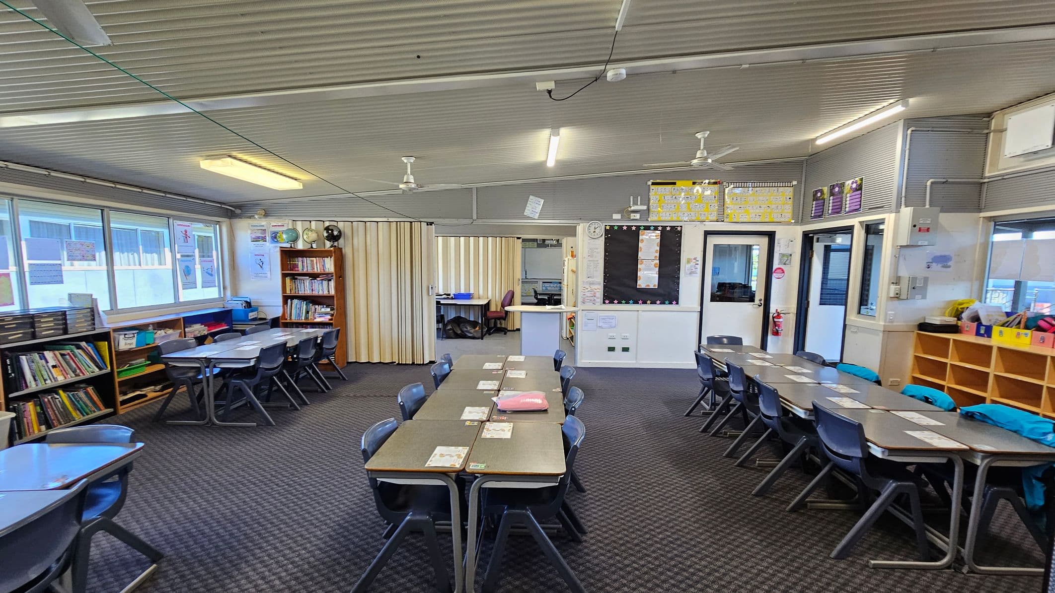A classroom with a grey carpeted floor and a corrugated ceiling. On the left, there are bookshelves filled with books and educational materials, along with a row of windows. In the centre, several tables are arranged with chairs around them, and some have papers and a red folder on top. The back wall has a partition, a clock, and a blackboard. On the right, there are more tables and chairs, a set of cubbies with colourful items, and two doors leading to other rooms. Ceiling fans and fluorescent lights are visible.