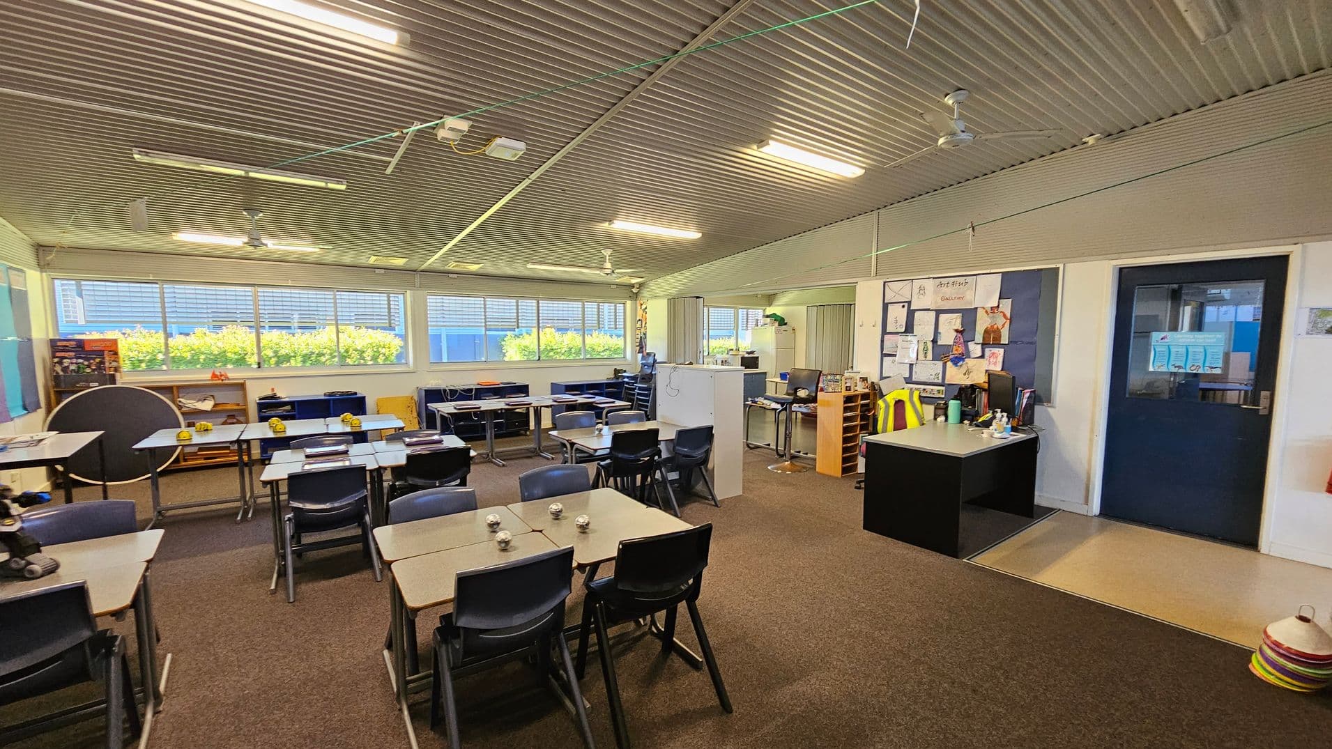 A classroom with a corrugated metal ceiling and fluorescent lights. On the left, there are several desks and chairs arranged in groups, with some educational materials on the tables. Large windows line the back wall, allowing natural light to illuminate the room. On the right, a teacher's desk is positioned near a bulletin board displaying various papers and artwork. A dark blue door is adjacent to the desk. The floor is carpeted, except for a small section of linoleum near the door.