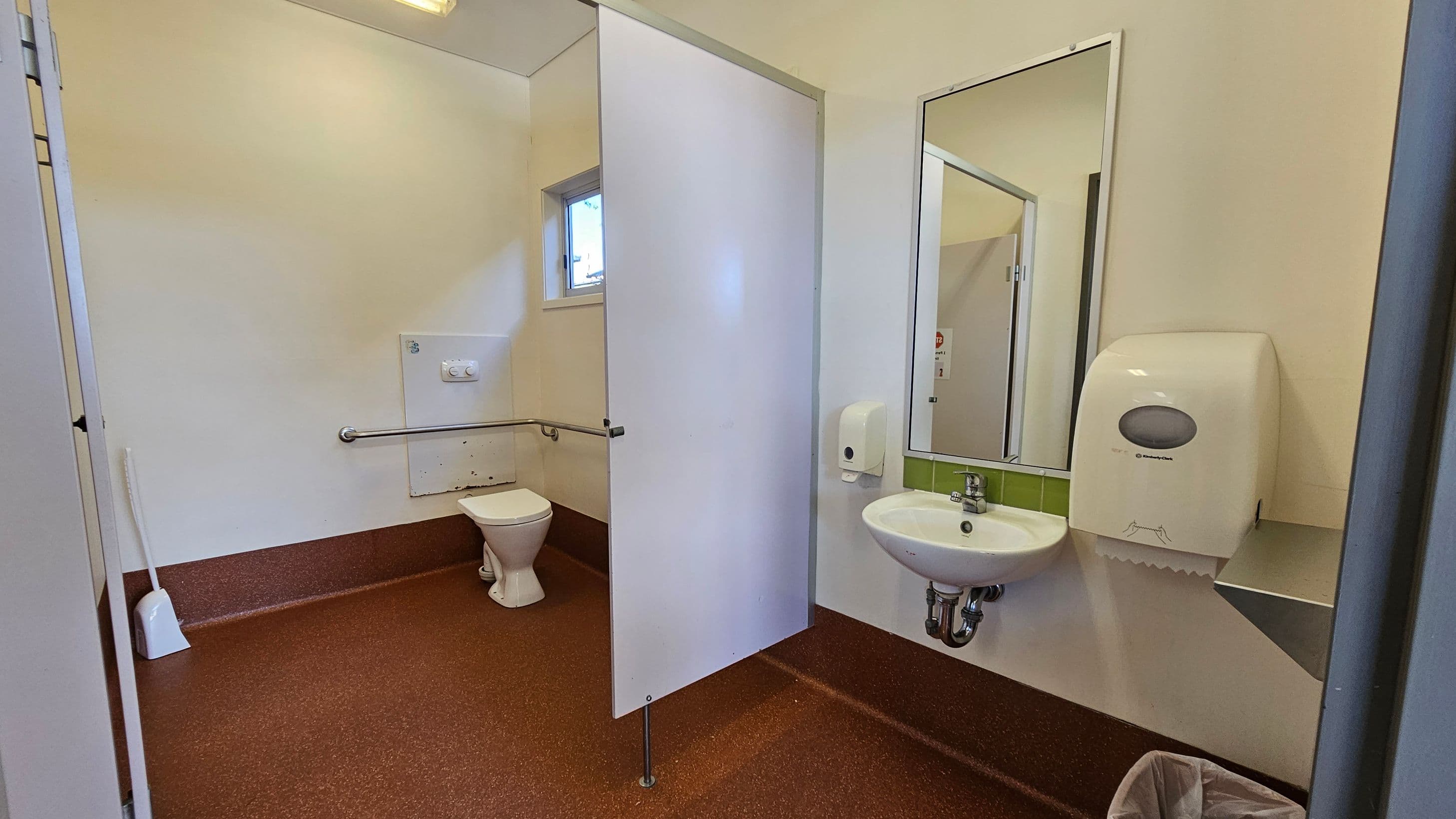 A public restroom with a red-brown speckled floor and cream walls. On the left, a toilet with a grab bar and a small window above. In the centre, a white partition partially separates the toilet area. On the right, a wall-mounted sink with a mirror above it, a soap dispenser, and a paper towel dispenser. A small waste bin is on the floor beneath the sink. The ceiling is plain with a light fixture.
