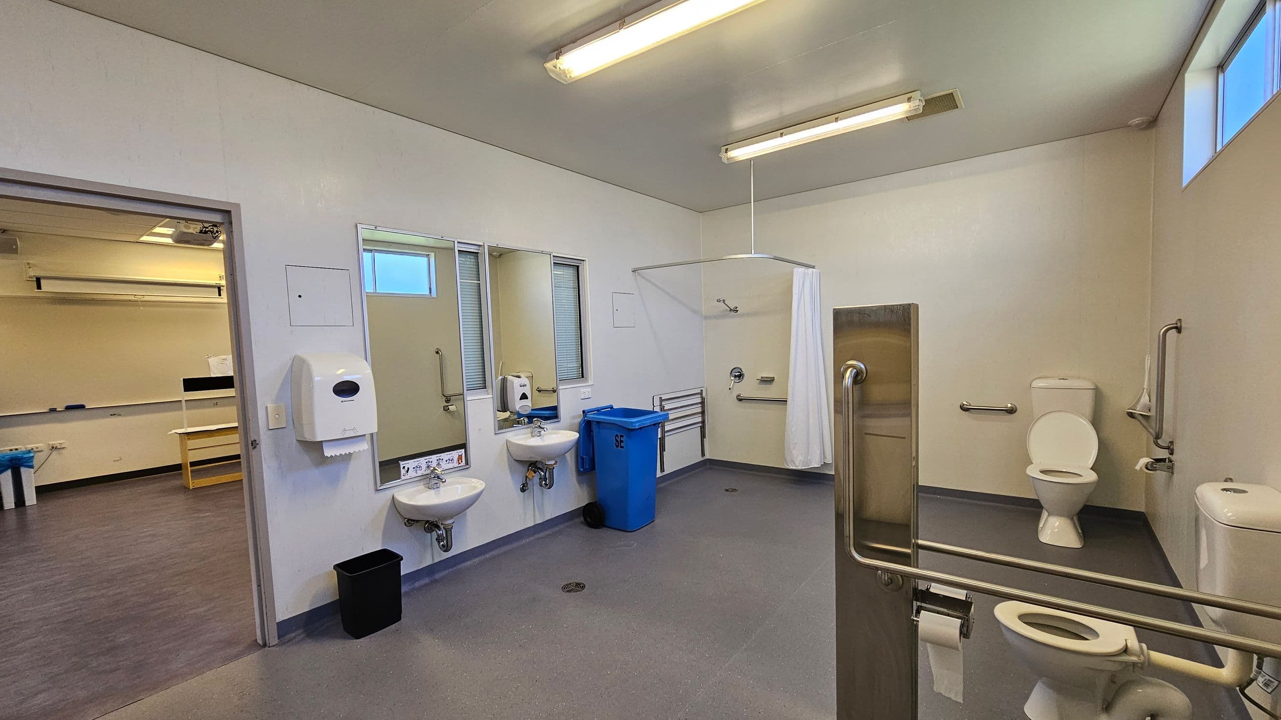 A spacious, accessible bathroom with a grey floor and white walls. On the left, there are two sinks with mirrors above them, a paper towel dispenser, and a soap dispenser. A blue bin is positioned nearby. In the middle, a shower area with a curtain and grab bars is visible. On the right, there are two toilets, each with grab bars for accessibility. The ceiling has fluorescent lights, and there are high windows allowing natural light in. An open door leads to an adjacent room with a purple floor.