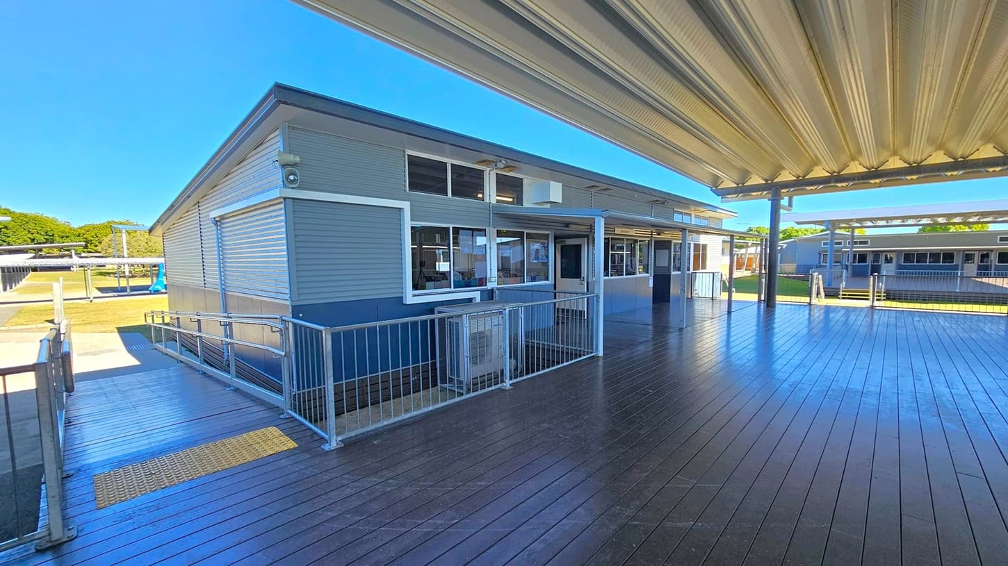 A modern building with metal siding and large windows is on the left. A wooden deck with a metal railing runs alongside it, leading to a ramp. The ceiling is covered with a corrugated metal roof. In the background, another similar building is visible, surrounded by a grassy area. The structures are connected by covered walkways.