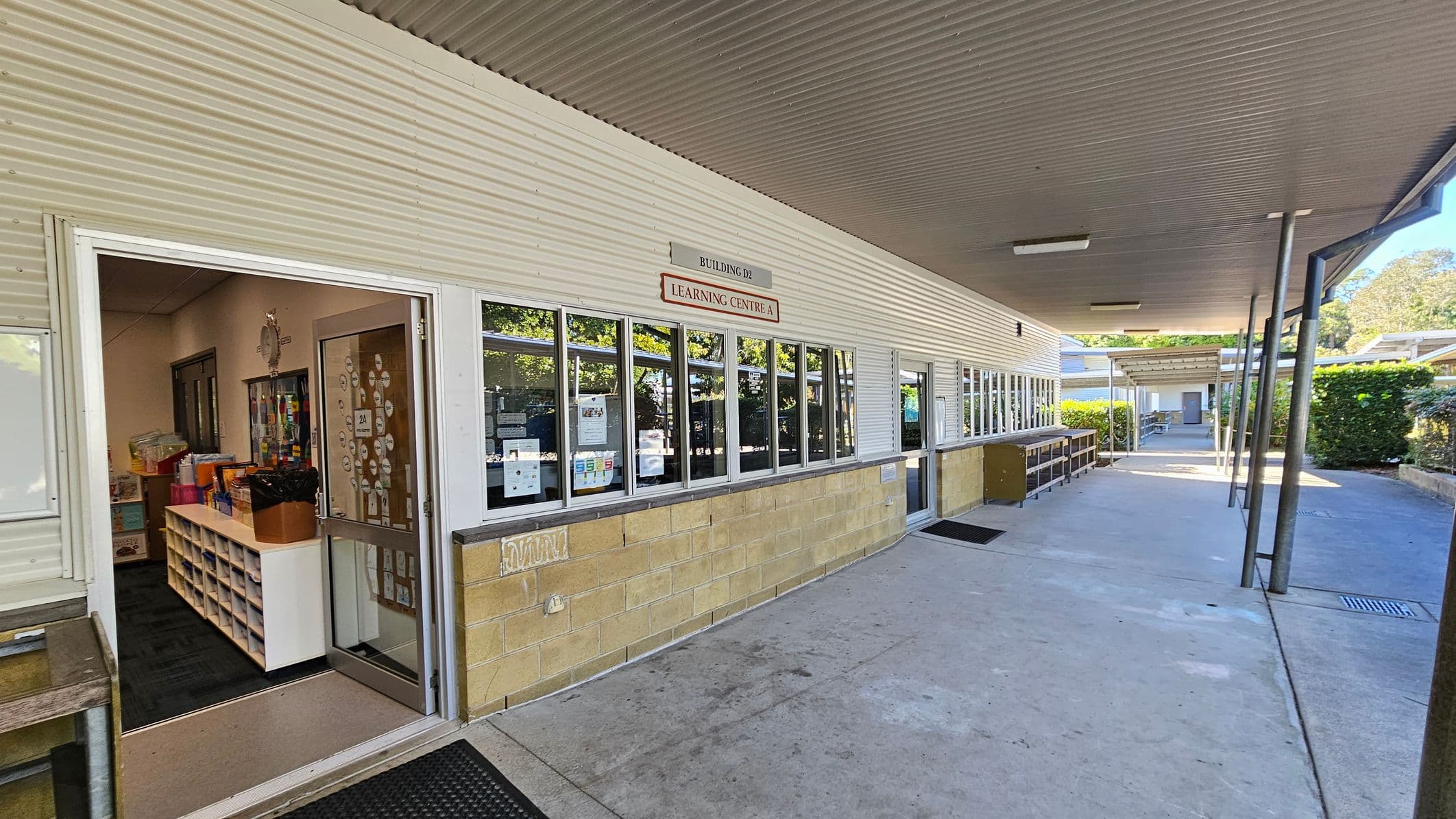 A covered walkway with a corrugated metal ceiling and fluorescent lights. On the left, there is a building with a sign reading "Building D2 Learning Centre A" above a row of windows. An open door reveals an interior with shelves and educational materials. The floor is concrete, and there are metal poles supporting the roof. In the background, there are more buildings and greenery, including hedges along the walkway.