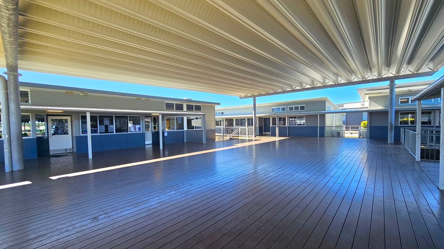 A spacious outdoor area with a wooden deck floor and a large, corrugated metal roof overhead. Surrounding the deck are several single-storey buildings with grey walls and white-framed windows and doors. The buildings have small overhangs and are connected by covered walkways. Metal railings are visible on the right side, and there are steps leading up to one of the buildings in the background. The area is well-lit, with sunlight casting shadows on the deck.