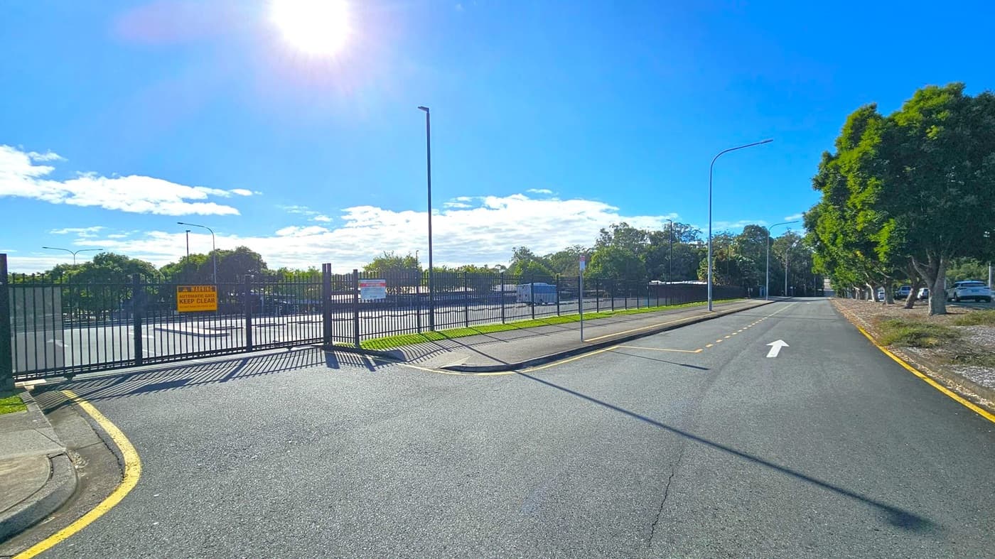 A road with a single white arrow pointing forward. On the left, there's a tall metal fence with a yellow warning sign about an automatic gate. Behind the fence, a parking area is visible. On the right, a row of trees lines the road, with a few parked cars beneath them. The road is bordered by a kerb and a narrow strip of grass. The sun is bright, casting shadows on the ground.