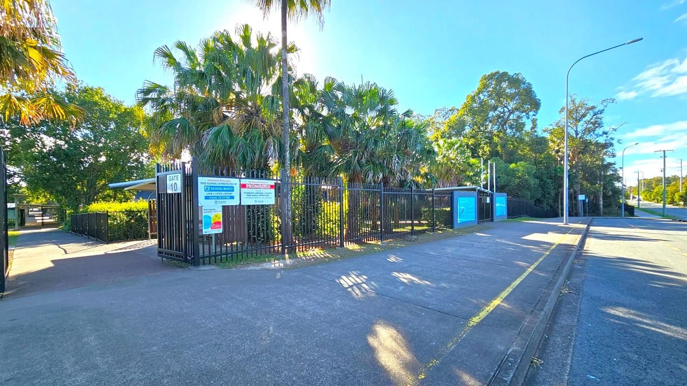 A gated entrance marked "Gate 10" with signs about school security and prohibited activities. Tall palm trees and dense greenery are behind the black metal fence. The pavement is wide, with a yellow line along the kerb. A streetlight and power lines run along the road on the right. The sky is clear and blue, casting shadows on the ground.