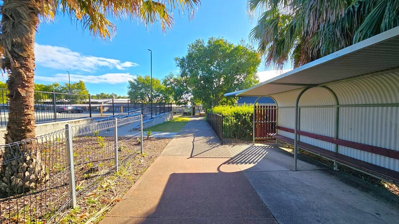A pathway runs through the centre, bordered by a metal fence on the left and a covered walkway on the right. The ground is a mix of concrete and reddish pavement. Palm trees and other greenery are visible on both sides, with a large tree in the background. A car park is visible to the left, separated by a fence. The covered area has a corrugated metal roof, casting shadows on the ground.