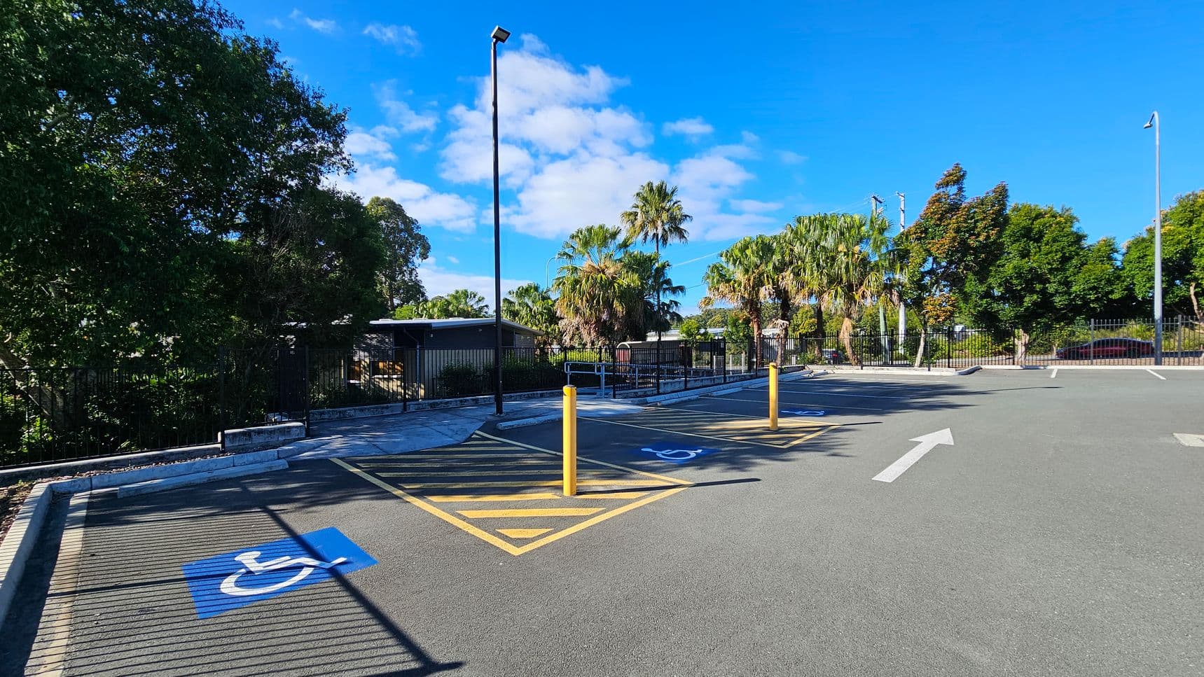 A parking area with designated disabled parking spaces marked by blue and white symbols on the asphalt. Yellow lines and bollards separate the spaces. On the left, there are large green trees, while the right side features more trees and a fence. In the background, palm trees and a building are visible. A white arrow is painted on the ground, indicating traffic direction. The area is well-lit by tall street lamps.