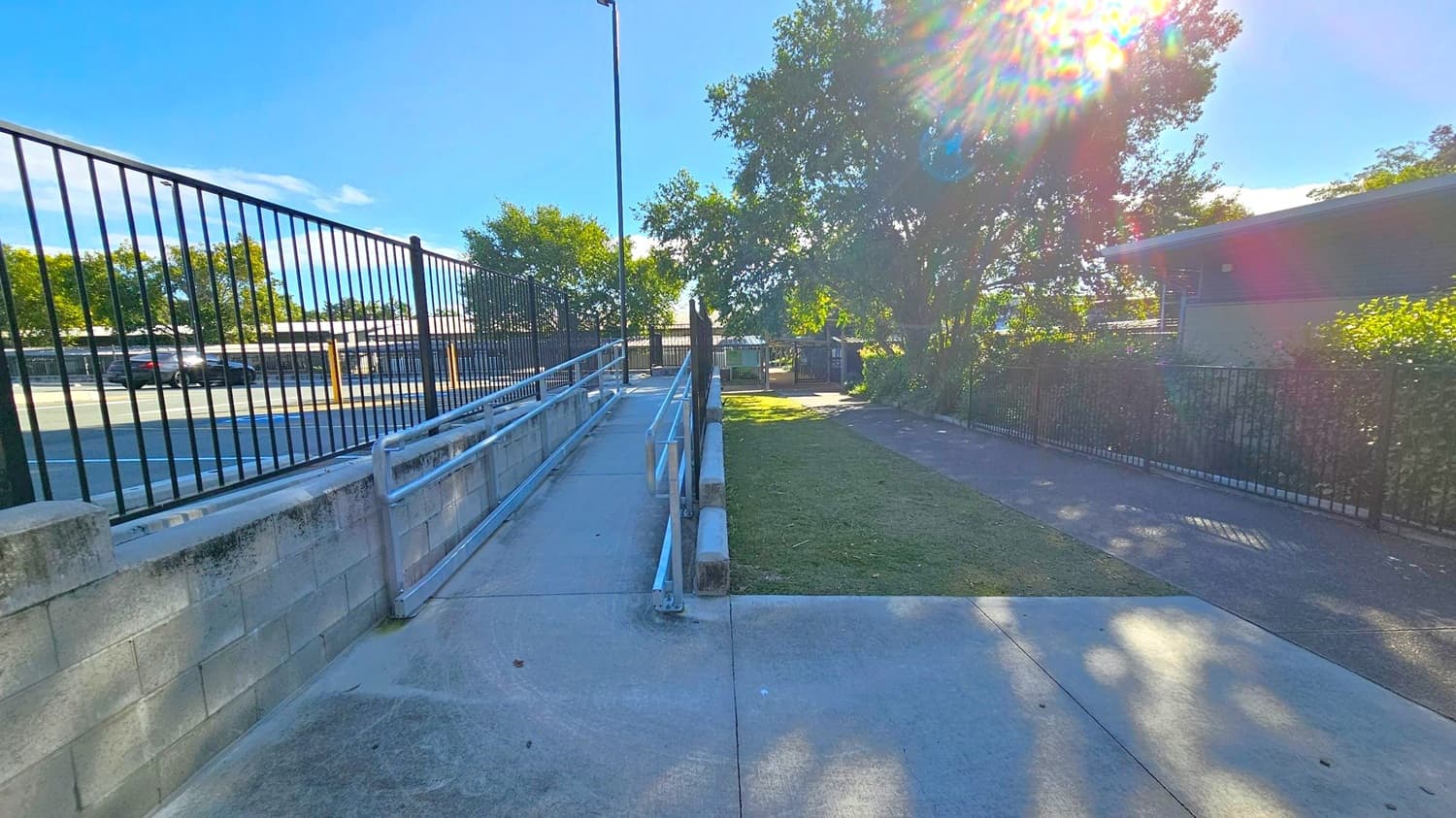 A concrete pathway with a metal railing runs diagonally from the foreground to the background on the left side, bordered by a low concrete wall and a black metal fence. On the right, there's a grassy area with a tree casting shadows, and a building partially visible behind more fencing. Sunlight creates a lens flare effect near the tree. A car park with a black car is visible through the fence on the left.