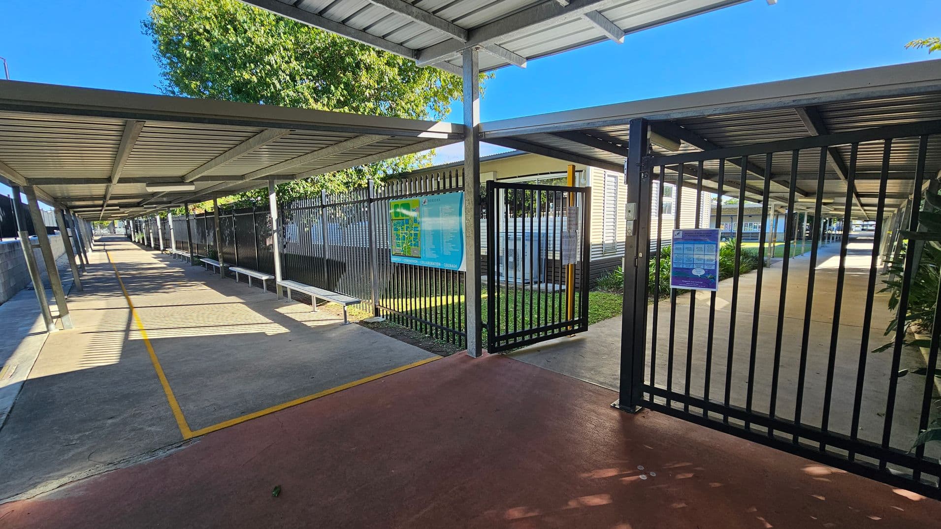 A covered walkway with a metal roof and support beams extends into the distance on both the left and right sides. The ground is paved with a yellow line marking the path. On the left, a metal fence with a map and benches runs alongside the walkway. On the right, a gate with a notice leads to another covered path. In the background, there are buildings with windows and some greenery. The ceiling is metal, matching the roof of the walkway.