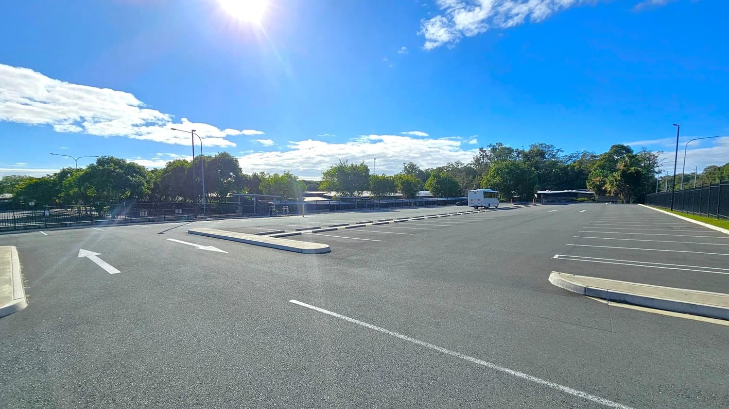 An empty car park with marked parking spaces and directional arrows on the asphalt. On the left, there are a few trees and a fence, while on the right, a line of trees and a fence run parallel to the parking area. In the background, a white vehicle is parked, and more trees are visible. The sun is shining brightly, casting shadows on the ground.