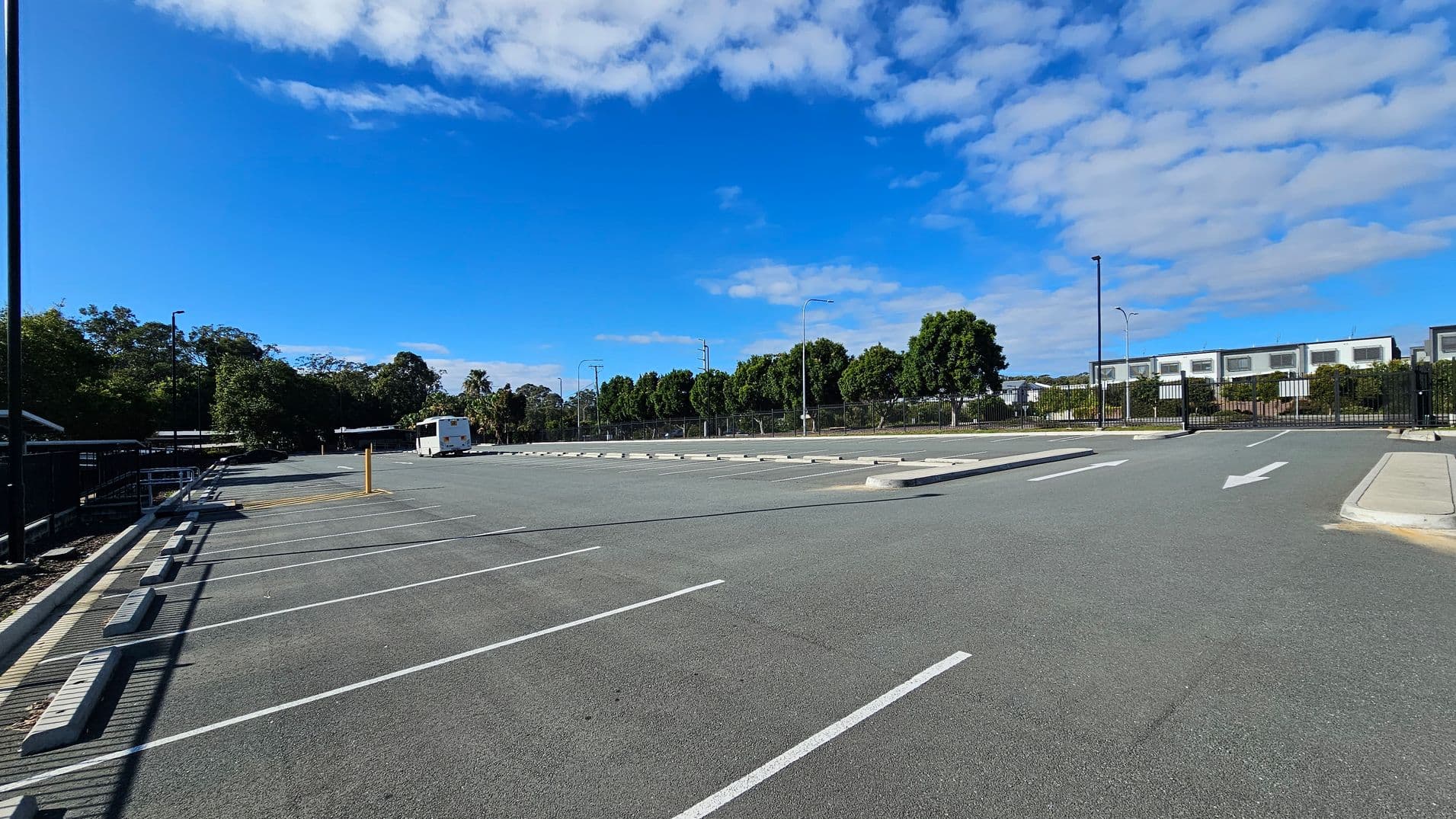 A large, empty car park with marked parking spaces on the left. A single white bus is parked in the distance. Trees line the background, with a fence and a row of modern buildings visible on the right. The ground is paved with asphalt, and there are several streetlights scattered throughout the area. White arrows on the ground indicate traffic direction.