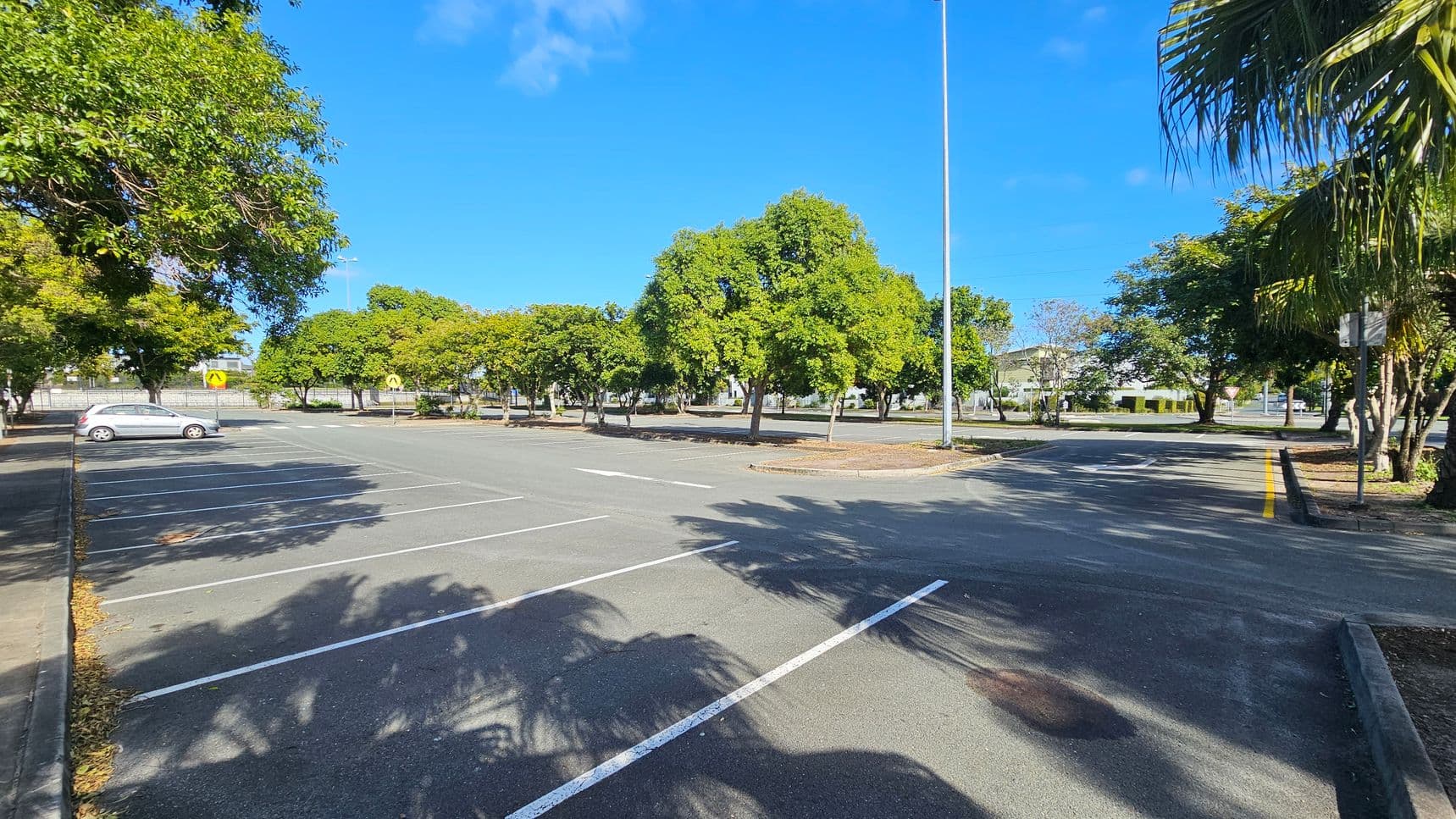 An empty car park with a single silver car parked on the left. The ground is marked with white parking lines and scattered leaves. Trees with green foliage line the background, casting shadows on the asphalt. A tall streetlight stands in the centre, and a yellow road sign is visible. On the right, a road with a yellow line runs alongside more trees. The sky is clear and blue.