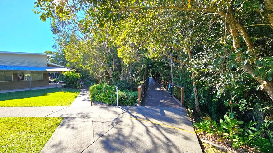 A pathway leads into a dense, leafy forest area on the right, with a wooden bridge visible. On the left, a building with a corrugated metal roof and windows is partially visible, surrounded by well-maintained grass and shrubs. The ground is paved, and sunlight filters through the trees, casting shadows. The sky is clear and blue.