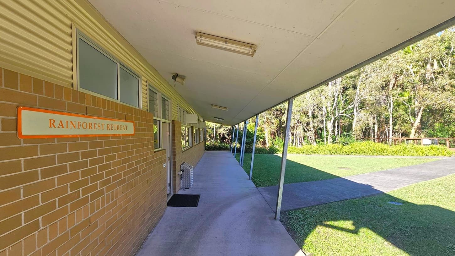 A covered walkway with a slanted roof and metal supports on the right. On the left, a brick wall with a sign reading "Rainforest Retreat" and several windows. The floor is concrete, leading to a grassy area with a path. In the background, there are trees and dense foliage. Ceiling lights are mounted along the roof.