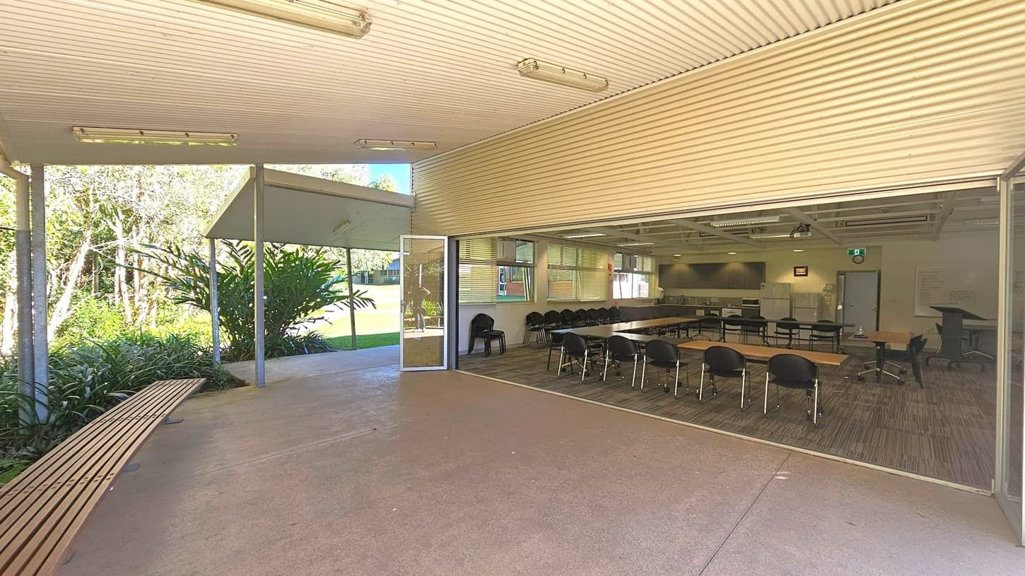 A covered outdoor area with a corrugated ceiling and fluorescent lights. On the left, there's a wooden bench and lush greenery with trees and plants. The right side features an open indoor space with tables and chairs arranged in rows, a projector, and a screen. The floor inside is carpeted with a patterned design, while the outdoor area has a concrete floor. A glass door separates the two spaces, and the indoor area has windows with blinds.
