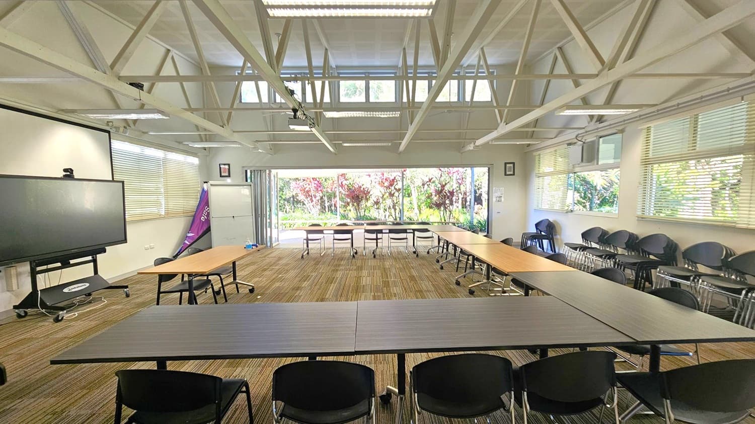 A conference room with a U-shaped arrangement of tables and chairs. On the left, there's a large screen on a stand and a window with blinds. The right side has stacked chairs and more windows with blinds. The back wall features large glass doors opening to a garden with red and green foliage. The ceiling has exposed beams and fluorescent lights, and the floor is carpeted with a striped pattern.