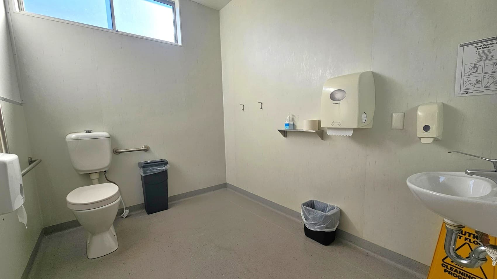 A public restroom with light green walls and a grey floor. On the left, there is a toilet with a grab bar and a black bin beside it. In the centre, a shelf holds a bottle and a roll of paper, with a paper towel dispenser above. On the right, a small sink with a soap dispenser is mounted on the wall, and a yellow caution sign is on the floor. A window at the top left lets in natural light.
