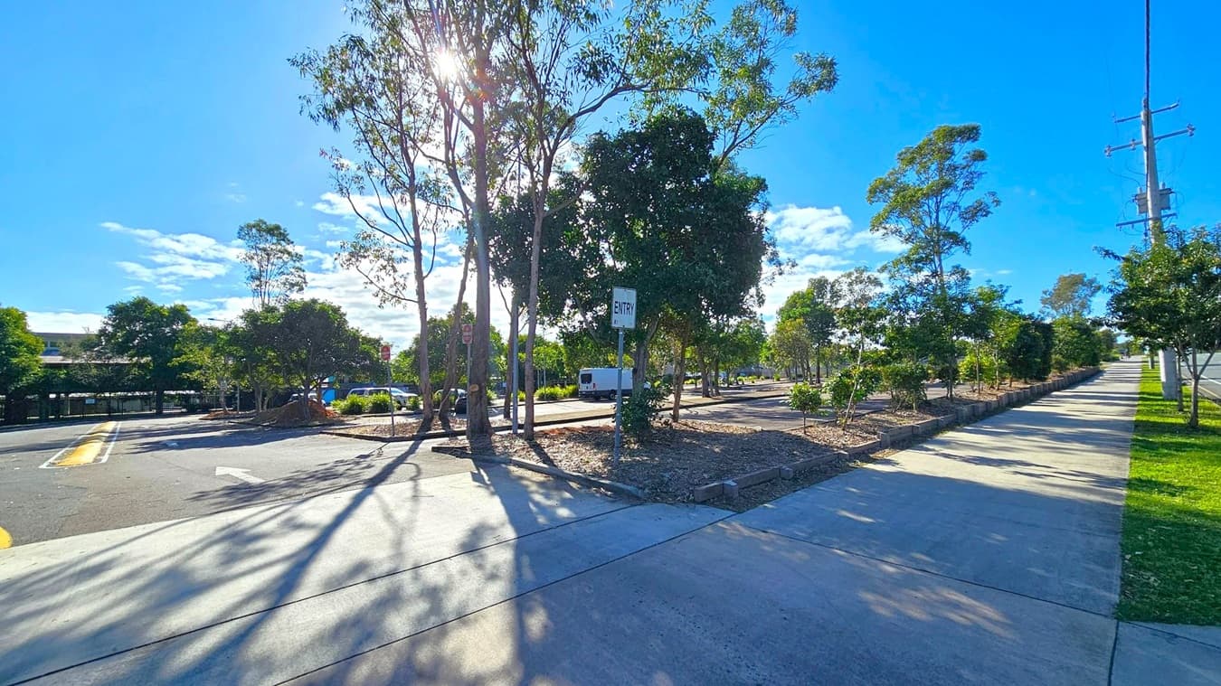 A street scene with a paved road and footpath. On the left, there are several trees casting shadows on the road, with a sign marked "ENTRY" visible. A white van is parked further down the road. On the right, a footpath runs alongside a row of trees and a grassy area. A utility pole is also visible on the right. The sun is shining brightly, creating long shadows on the ground.