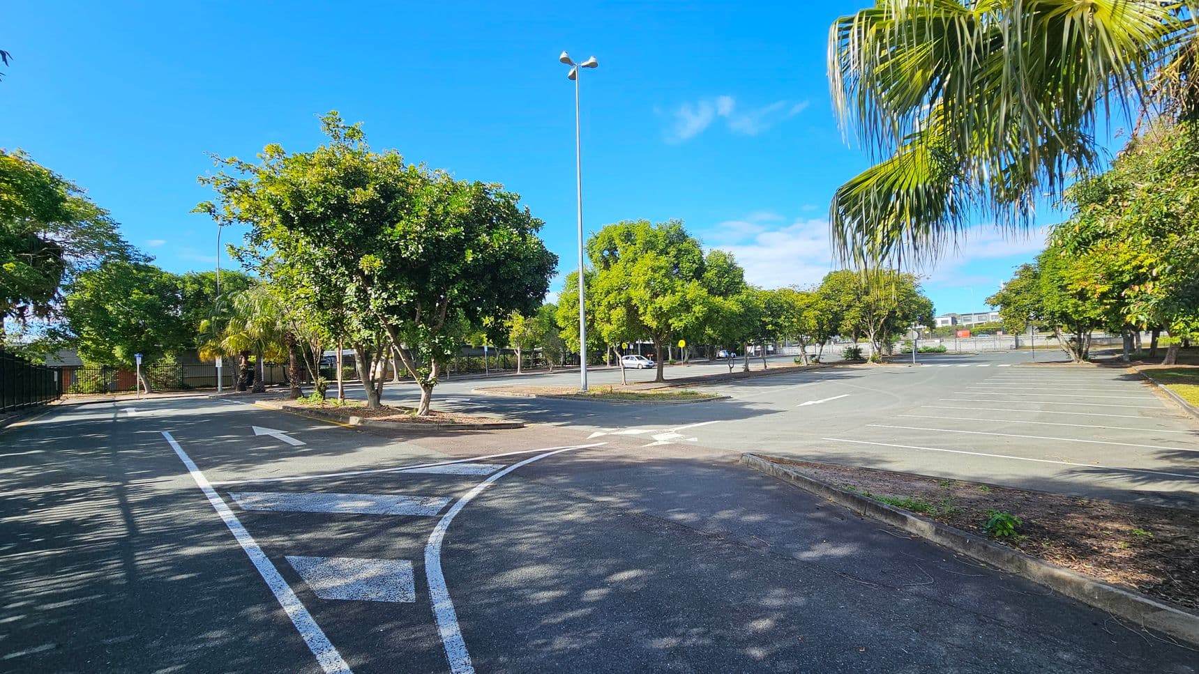 A nearly empty car park with marked parking spaces and a few scattered trees providing shade. A tall streetlight stands in the centre. The ground is asphalt with white directional arrows painted on it. On the left, a curved road leads out of the car park, bordered by more trees and a fence. On the right, palm fronds hang down, and more trees line the perimeter. In the background, a building is visible beyond the fence.