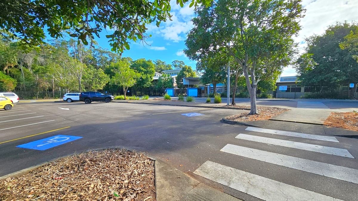 A car park with several vehicles, including a yellow car on the left and a black SUV in the middle. The foreground features a pedestrian crossing and a disabled parking space marked in blue. Trees with green foliage are scattered throughout, providing shade. In the background, a building with a modern design is partially visible, surrounded by more trees and greenery. The ground is covered with asphalt, and there are patches of dry leaves near the edges.