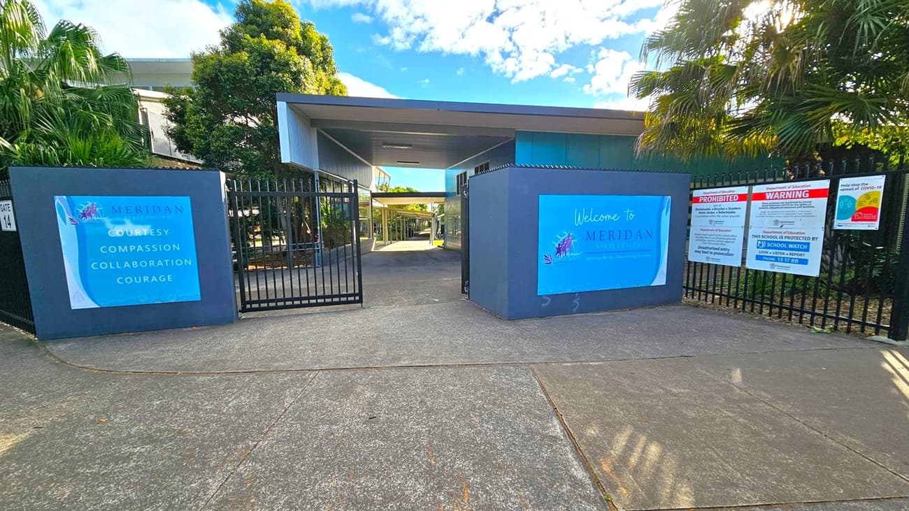 Entrance to a school with a wide concrete path leading to a gate. On the left, a large sign displays the words "Courtesy, Compassion, Collaboration, Courage" with the name "Meridan". On the right, another sign reads "Welcome to Meridan State College" alongside various warning and informational signs. Lush green trees and plants are visible on both sides, with a modern building structure in the background. The sky is partly cloudy.