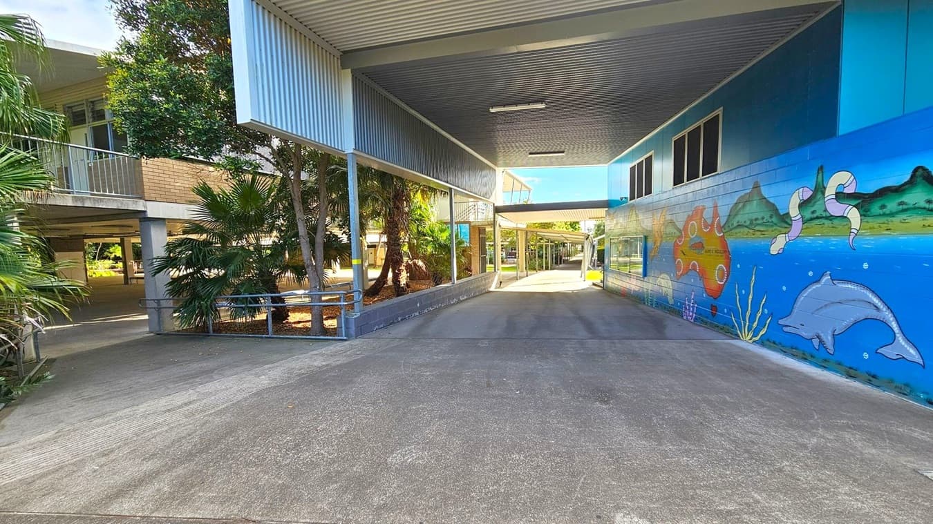 A covered walkway with a corrugated metal ceiling. On the right, a vibrant mural features a dolphin, sea creatures, and a map of Australia. The left side has a building with a balcony and lush greenery, including palm trees. The ground is paved, and the area is well-lit with natural light.