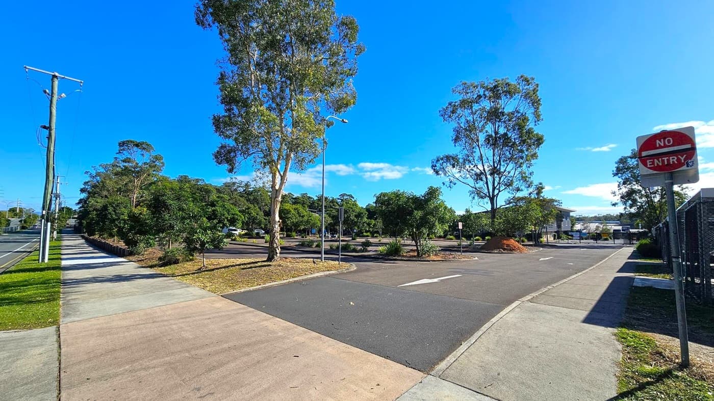 A paved footpath runs along the left side, bordered by grass and a row of utility poles. In the centre, a road leads into a parking area with several trees and a streetlamp. On the right, a "No Entry" sign is visible near the entrance of a fenced area. In the background, there are more trees and a building. The scene is well-lit, suggesting a sunny day.