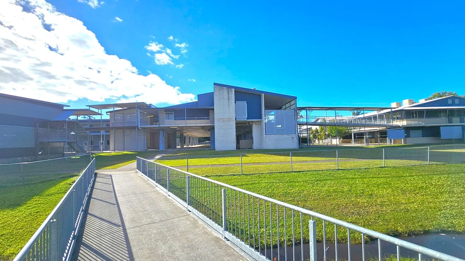 A modern building with a flat roof and large windows is in the centre, surrounded by a grassy area. A concrete path with metal railings leads towards the building. On the left, there is another structure with a staircase. To the right, additional buildings are connected by a covered walkway. The sky is clear with some clouds visible.