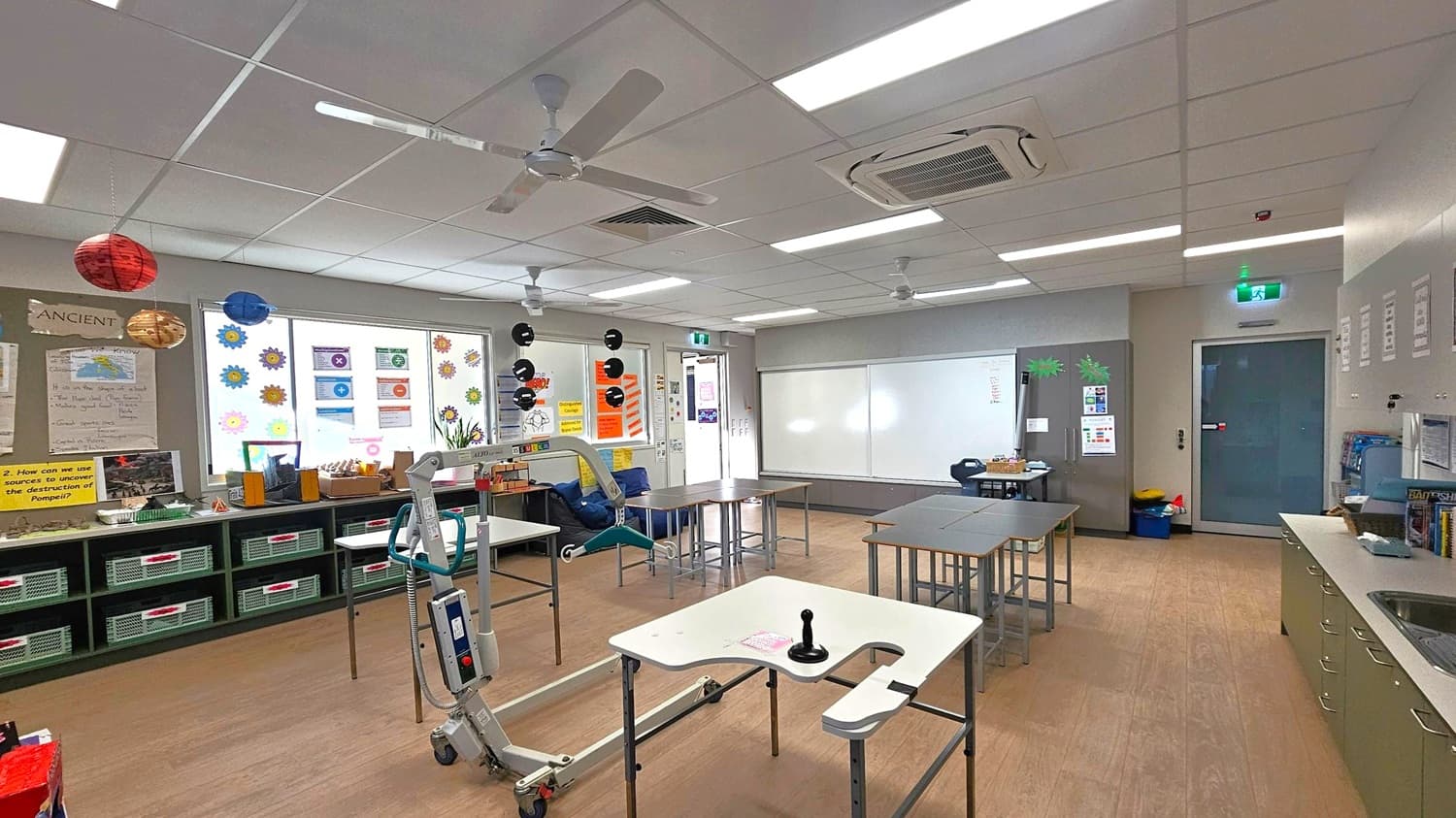 A classroom with a wooden floor and a white ceiling featuring fluorescent lights and ceiling fans. On the left, there are shelves with baskets and educational posters, including one about Pompeii. Colourful paper planets hang from the ceiling. In the centre, there are tables and a mobility aid. The back wall has a large whiteboard and a door with posters. On the right, a counter with a sink and storage cabinets. The room is well-lit and organised.
