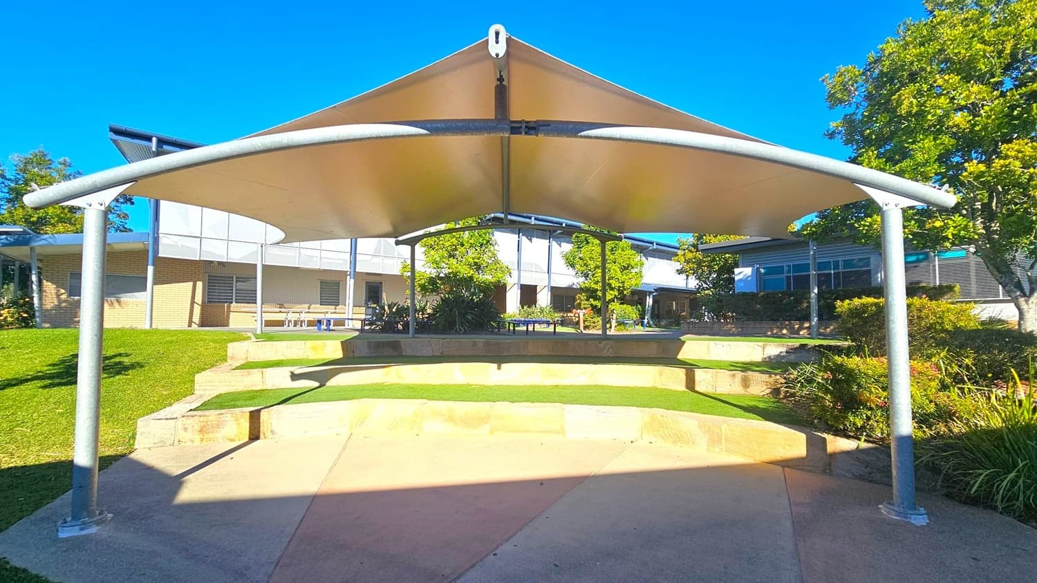A shaded outdoor area with a white fabric canopy supported by metal poles. Beneath the canopy, there are tiered stone seating steps with patches of grass. In the background, a building with brick and metal siding is visible, along with several windows. Trees and shrubs are scattered around the area, providing greenery. The ground is a mix of concrete and grass, and the sky is clear and blue.