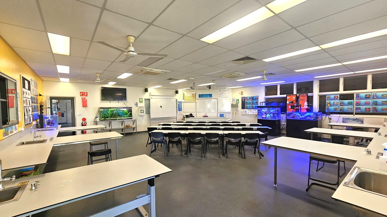 A classroom with a focus on science. On the left, there are long tables with sinks and taps. The back wall features a large fish tank and a wall-mounted TV. In the centre, rows of desks and chairs face a whiteboard and projector screen. The right side has more fish tanks and educational posters. The ceiling is fitted with fluorescent lights and ceiling fans. The floor is dark and speckled.