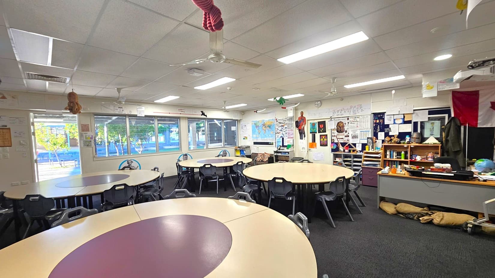 A classroom with round tables and black chairs arranged in the centre. The left side has large windows with a view of trees outside. The right side features a wall with educational posters, a world map, and a shelf filled with books and educational materials. The ceiling has fluorescent lights and a fan. The floor is carpeted in dark grey. Various decorations hang from the ceiling, and there are colourful items on the walls.