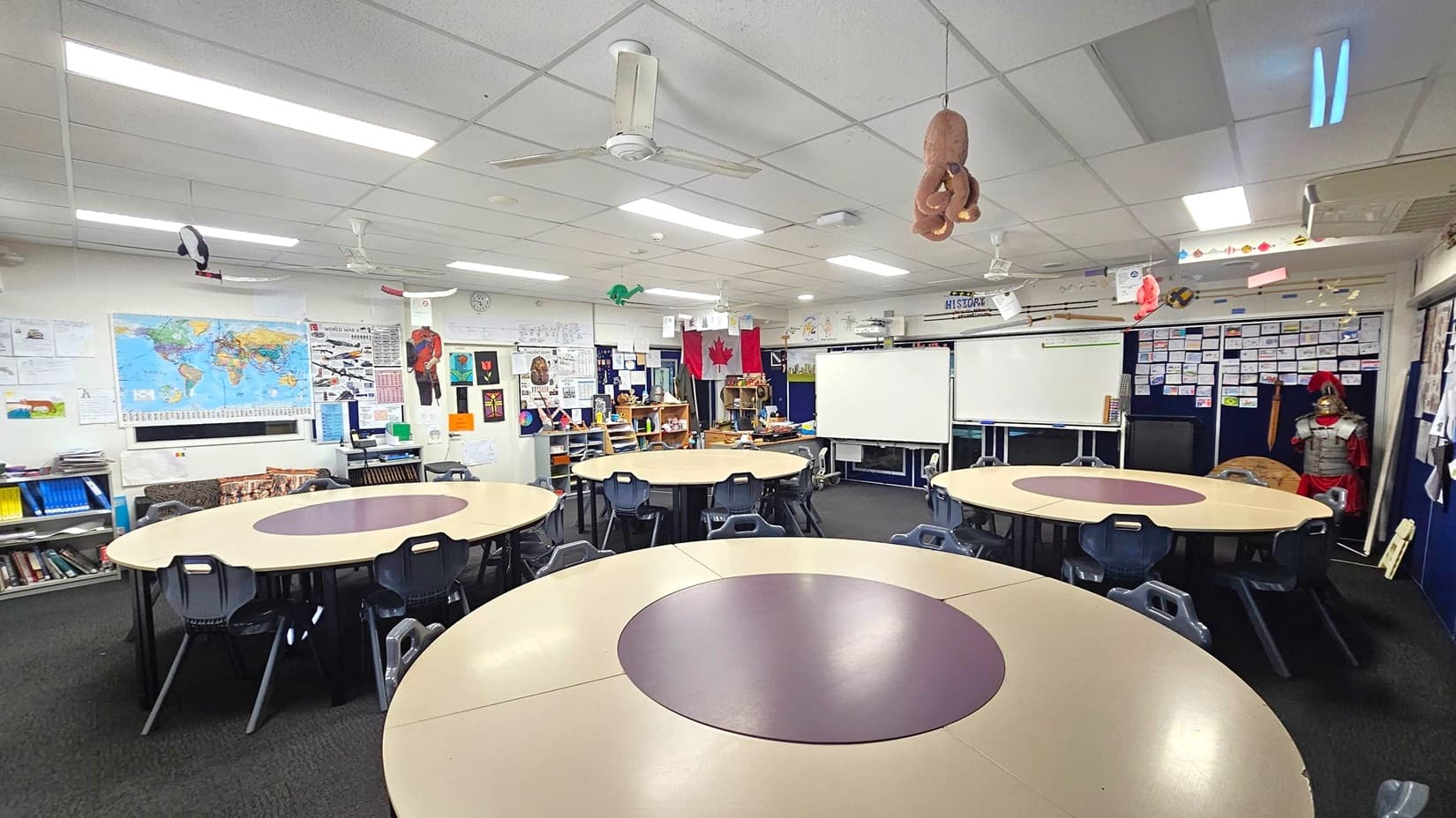 Classroom with circular tables and black chairs arranged in the centre. The walls are decorated with educational posters, including a world map and various artworks. A whiteboard is positioned at the back. Shelves with books and educational materials line the left side. A mannequin dressed in armour stands to the right. The ceiling has fluorescent lights and hanging decorations, including a plush toy. The floor is carpeted in dark grey.