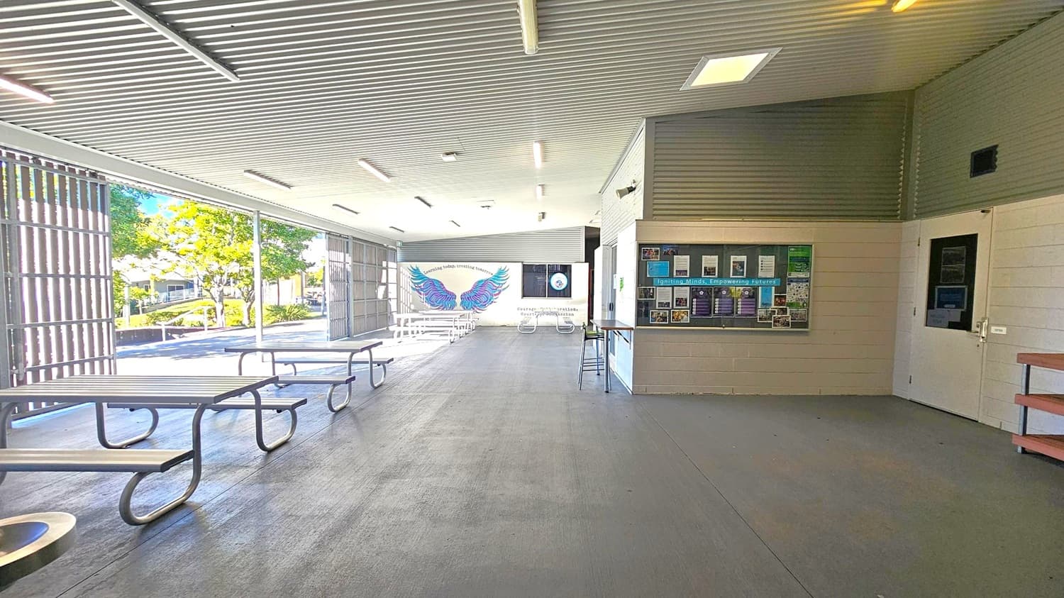 An open-air space with a corrugated metal ceiling and concrete floor. On the left, there are picnic tables with metal frames and wooden tops. The background features a wall with a colourful mural of wings and some text. On the right, a bulletin board displays various notices and a door is visible. The area is well-lit with fluorescent lights, and outside, trees and a pathway can be seen through the open side.