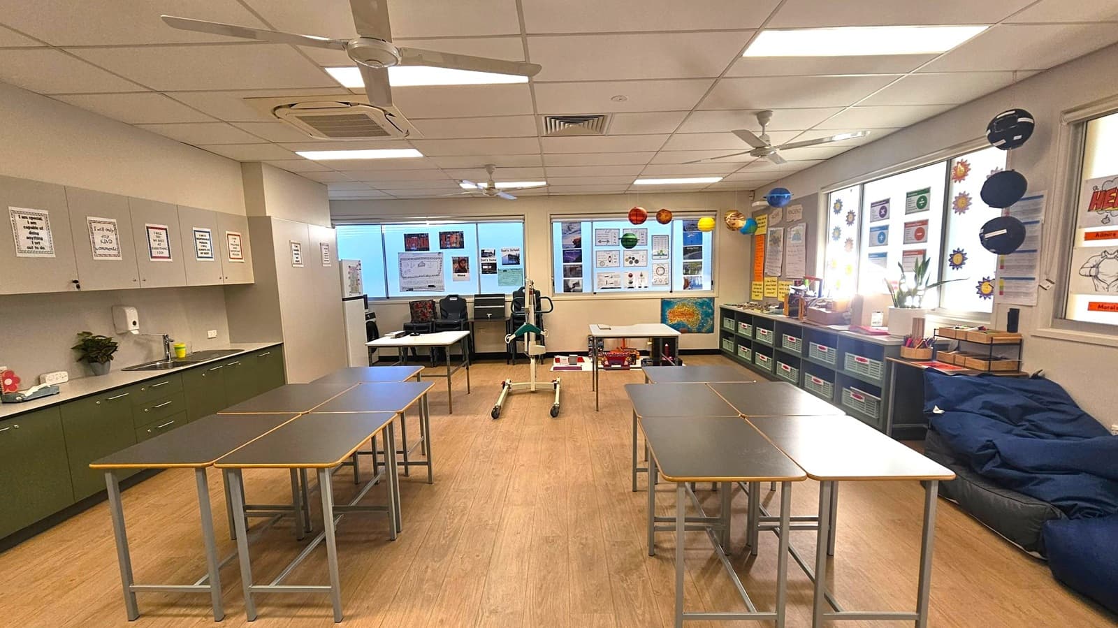 A classroom with a wooden floor and a ceiling with fluorescent lights and ceiling fans. On the left, there are green cabinets with educational posters above and a sink. In the middle, several tables are arranged in a U-shape. The back wall features windows with educational posters and hanging models of planets. On the right, there are storage bins, more educational materials, and a blue bean bag chair.