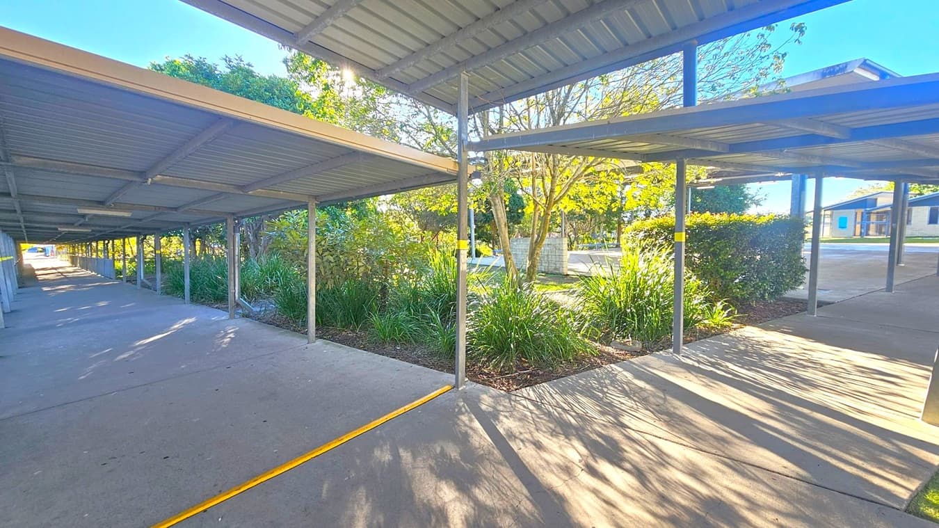 Covered walkways with metal roofs and supporting poles run parallel on the left and right. The ground is concrete with a yellow line marking. In the middle, there are landscaped garden beds with green plants and small trees. Sunlight filters through the foliage, casting shadows on the ground. In the background, a building with blue windows is visible, and more trees and shrubs are present. The ceiling is made of corrugated metal.