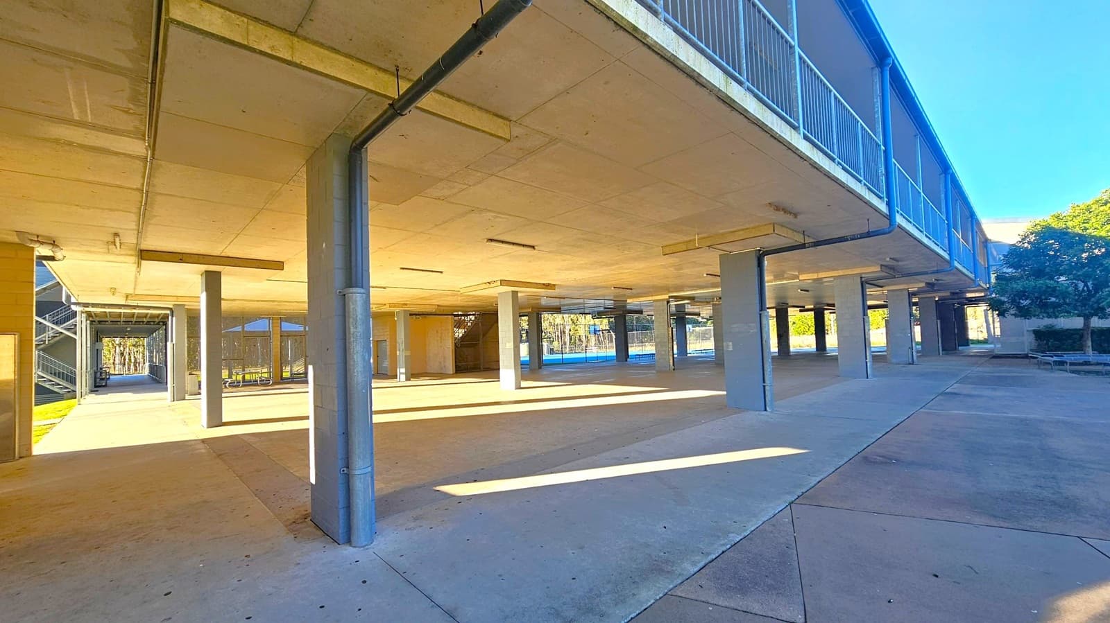 Concrete structure with multiple pillars supporting an upper level. On the left, a walkway leads to a staircase. The middle area is open, with shadows cast by the pillars. On the right, a railing lines the upper level, and a tree is visible near the edge. The floor is a mix of concrete and paved surfaces, and the ceiling is flat with visible beams and lighting fixtures.