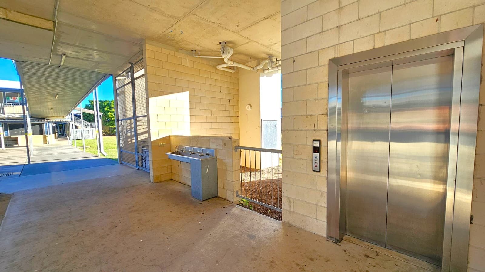 An outdoor area with a concrete floor and a ceiling with visible pipes. On the right, there is a stainless steel lift with a control panel beside it. In the middle, a small railing separates the lift area from a section with a drinking fountain attached to a brick wall. On the left, a covered walkway leads to a building in the background, with a grassy area visible beyond.