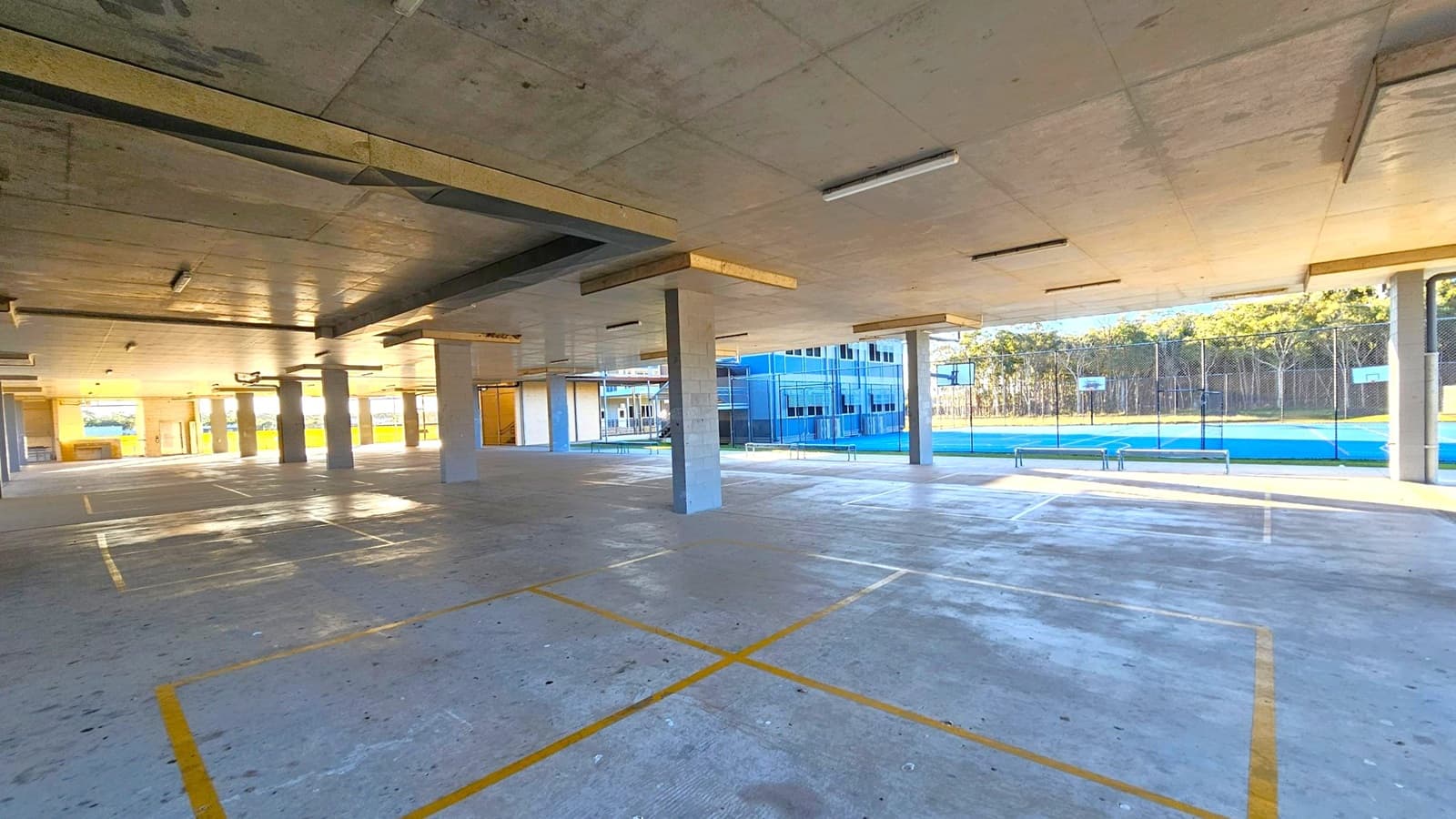 A spacious indoor area with a concrete floor marked with yellow lines, possibly for parking or sports. The ceiling is also concrete with fluorescent lights. On the right, there is a view of an outdoor basketball court with blue flooring, surrounded by a wire fence. In the background, there are trees and a building with large windows. The area is supported by several square columns.