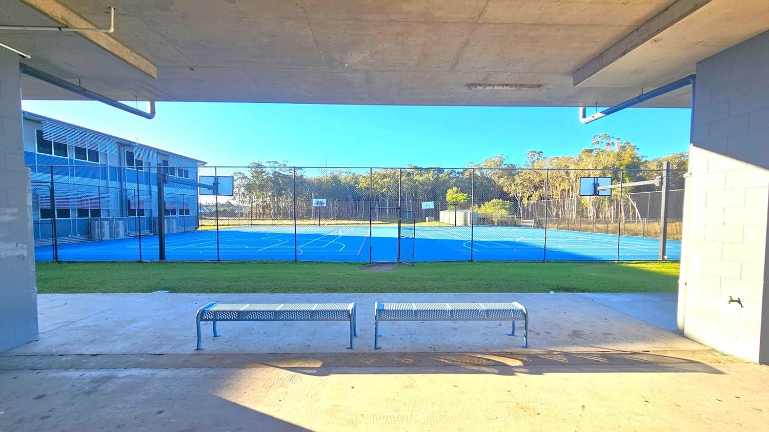 A view from under a concrete shelter with a ceiling, looking out towards a blue sports court with basketball hoops. In the foreground, there are two metal benches on a concrete floor. The court is enclosed by a chain-link fence. To the left, a multi-storey building with louvred windows is visible. In the background, there are trees and a clear blue sky.
