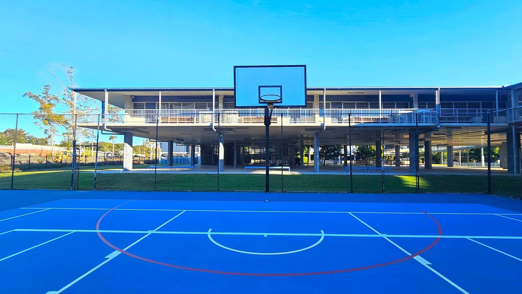 A blue outdoor basketball court with white and red lines is in the foreground. A basketball hoop stands at the centre. Behind the court, a two-storey building is elevated on pillars, with a grassy area underneath. The building has a balcony with railings. To the left, a tree is visible, and a fence surrounds the court. The sky is clear and blue.