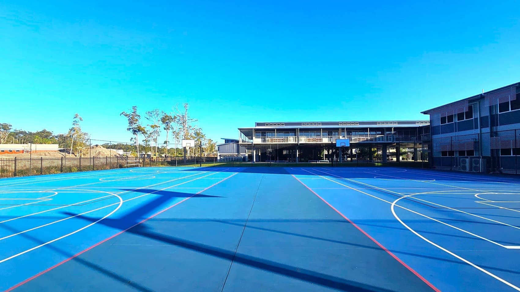A blue outdoor basketball court with white and red lines is in the foreground. Two basketball hoops are visible, one on the left and one on the right. In the background, there are two modern buildings with large windows. To the left, there are trees and a fenced area with some construction materials. The court is well-lit by sunlight, casting shadows on the surface.