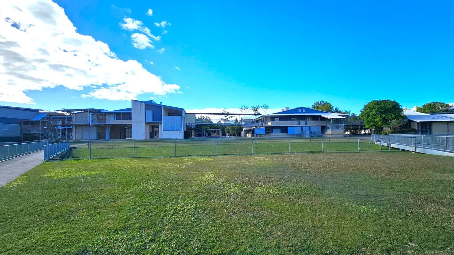 A grassy field in the foreground with a metal fence running across it. In the background, there are several modern, two-storey buildings with flat roofs and large windows. A paved path with a railing is on the left, leading towards the buildings. Trees are scattered around the area, and the sky is clear with some clouds on the left.