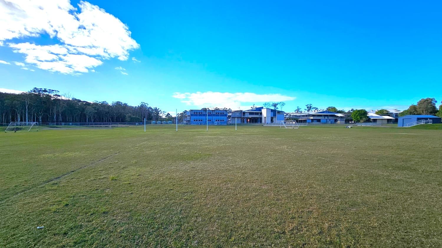 A grassy sports field with several goalposts is in the foreground. In the background, there are multiple modern buildings with flat roofs, likely part of a school or sports complex. To the left, a line of trees borders the field. The sky is clear with a few clouds.