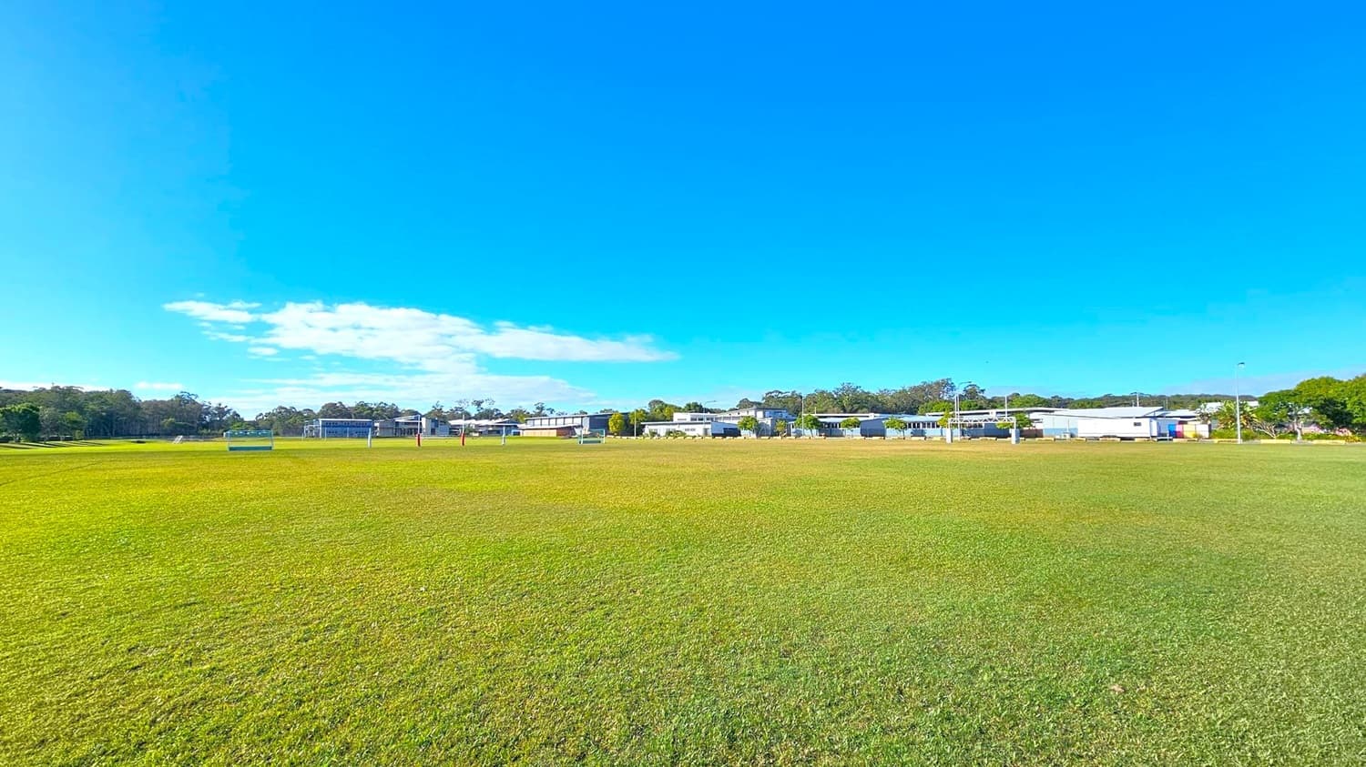 A wide grassy field stretches across the foreground, with a few goalposts and signs scattered throughout. In the background, there are several low buildings, possibly part of a school or sports complex, surrounded by trees. The sky is clear and blue, with a few clouds visible on the left side.