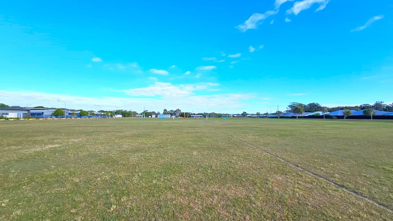 A wide, open grassy field stretches across the foreground, with patches of brown and green. In the background, there are several low buildings and houses, some with blue roofs, lined along the horizon. Trees are interspersed between the structures, and a few streetlights are visible. The sky is clear and blue, with a few scattered clouds.