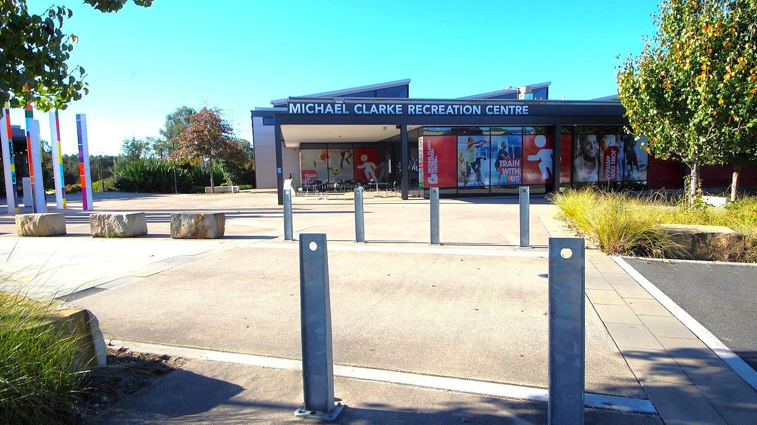 The view metres away from the front of Michael Clarke Recreation Centre. Several steel bollards with a round hole at the top and sandstone blocks can be seen in the entryway leading to the venue's entrance. Surrounding the venue are trees and bushes with stunning diversity. The venue's name can be visibly seen at the top of the building.